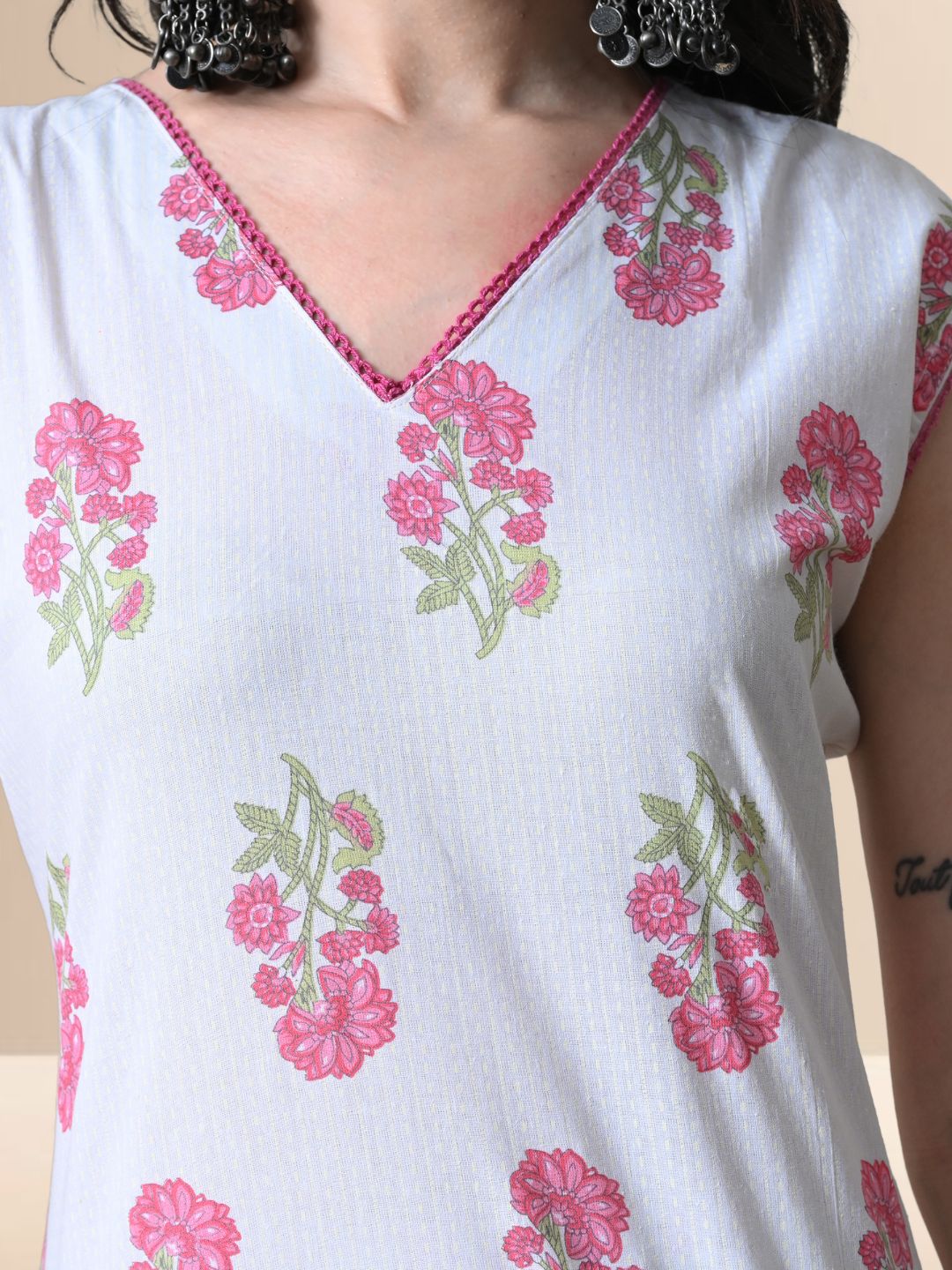 A woman wearing a white sleeveless kurta with pink floral prints and V-neck lace detail, paired with matching wide-leg floral pants from The Desi Cloth.