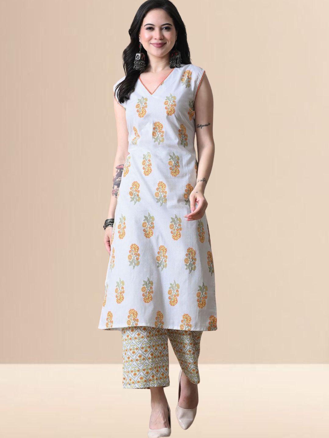 A woman wearing a sleeveless white cotton kurta with yellow and orange floral prints and a lace-trimmed V-neck, paired with matching printed wide-leg pants from The Desi Cloth.
